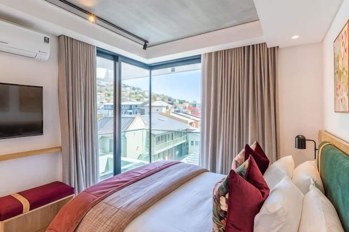 A hotel room with a large bed, a TV mounted on the wall, and a view of buildings through large windows.