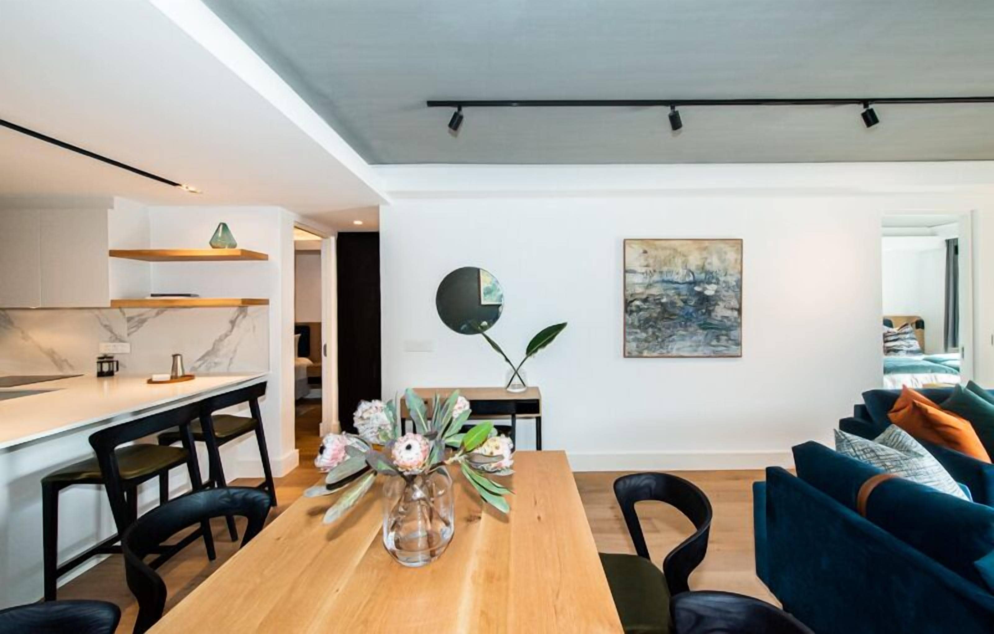 En moderne stue med et træspisebord, sorte stole og en blå sofa. Der er et køkkenområde med hvide skabe og en marmorbordplade. En vase med blomster står på bordet, og et maleri hænger på væggen.