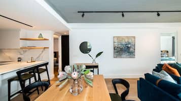 A modern living room with a wooden dining table, black chairs, and a blue sofa. There is a kitchen area with white cabinets and a marble countertop. A vase with flowers is on the table, and a painting is on the wall.