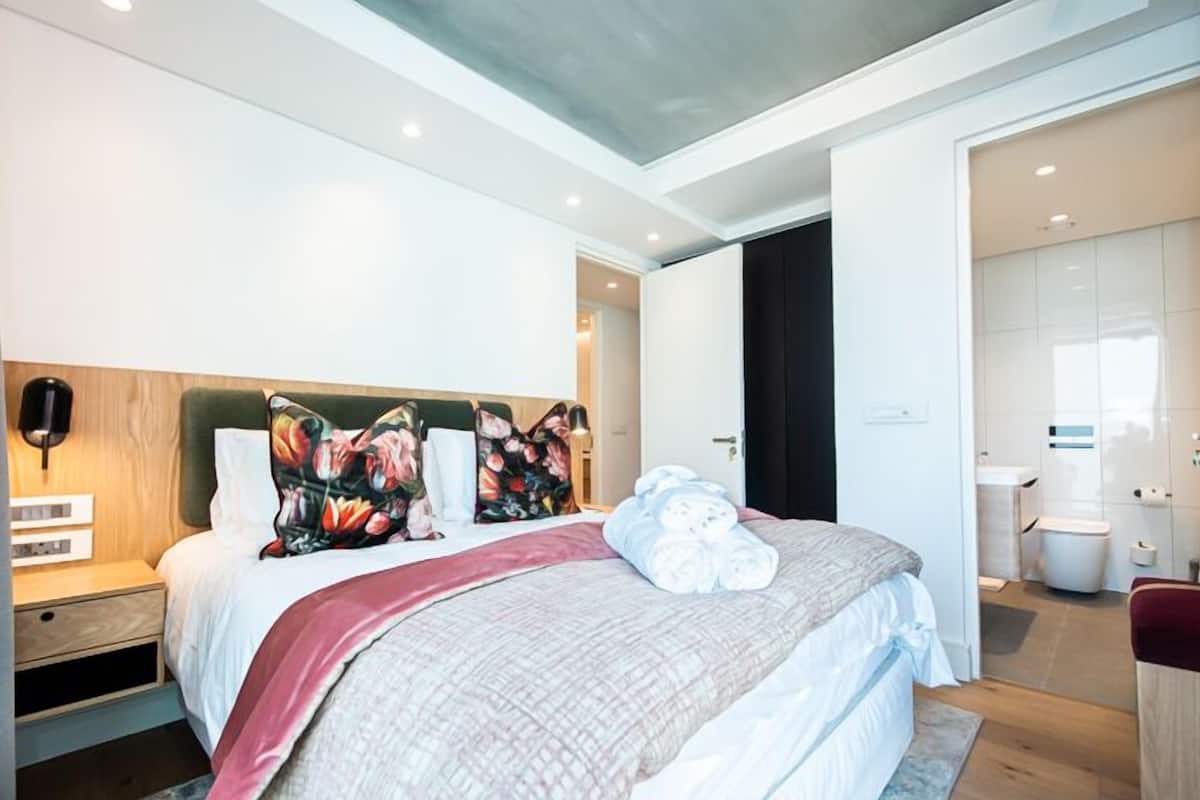 A modern hotel room with a large bed, a wooden headboard, a nightstand, a wall-mounted lamp, and a bathroom visible through an open door.