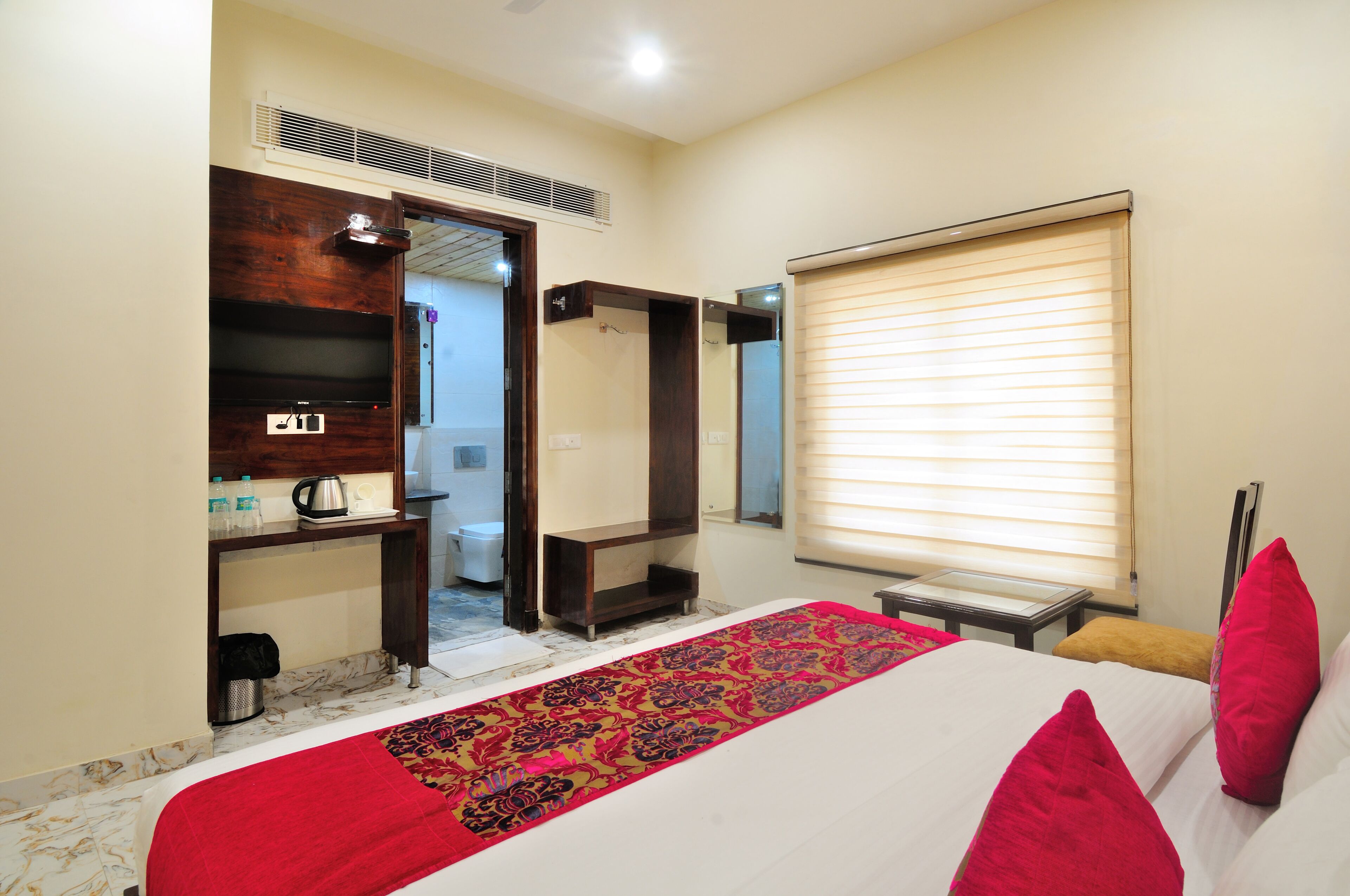 A hotel room with a bed, a red and black patterned runner, a wooden shelf with a TV and bottles, a window with blinds, and a door leading to a bathroom.
