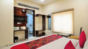 A hotel room with a bed, a red and black patterned runner, a wooden shelf with a TV and bottles, a window with blinds, and a door leading to a bathroom.