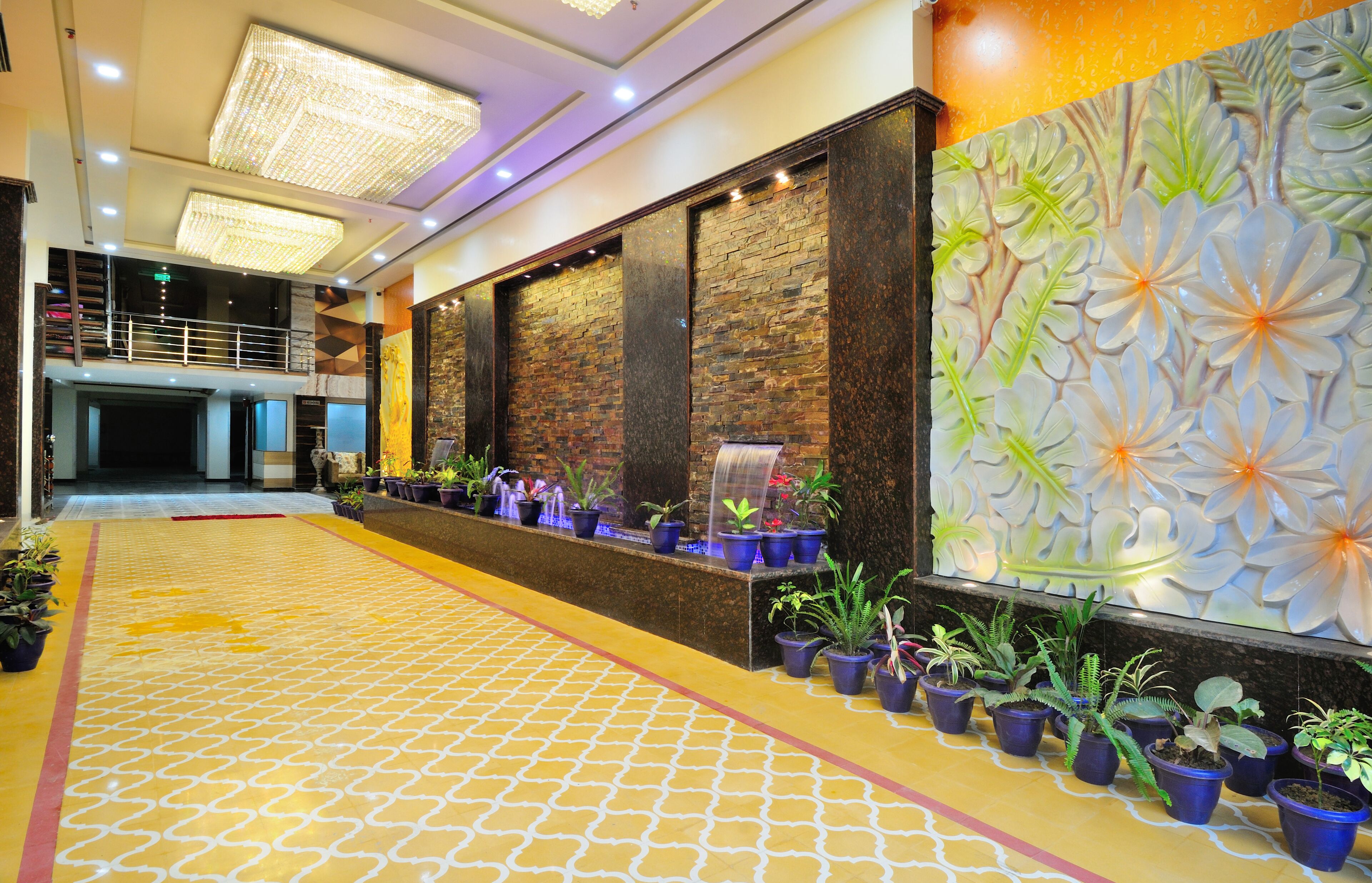 A hotel lobby with a patterned floor, potted plants, a water feature, and a decorative wall with floral designs.