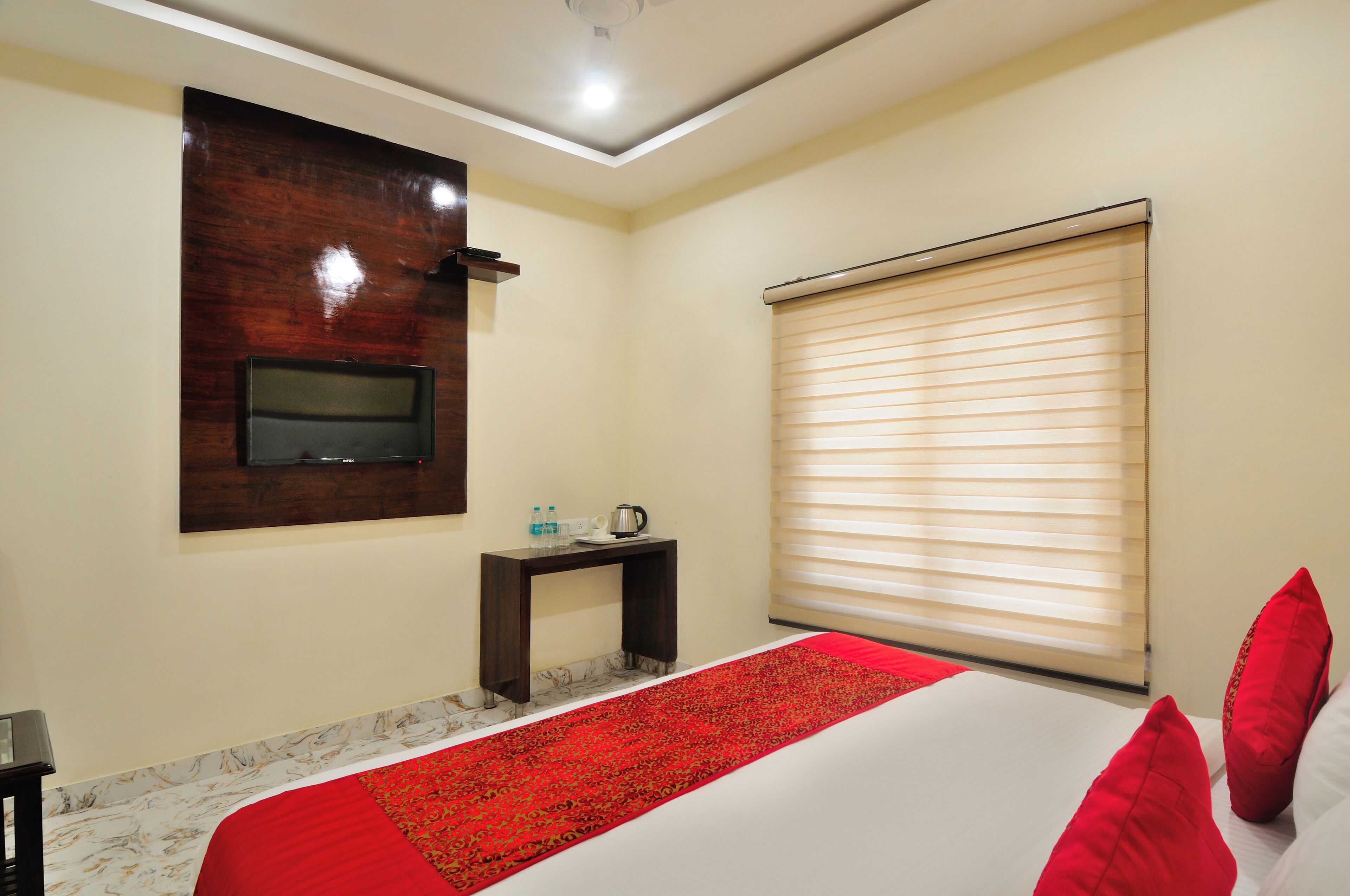 Habitación de hotel con cama, televisor montado en la pared, mesita con hervidor y ventana con persianas.