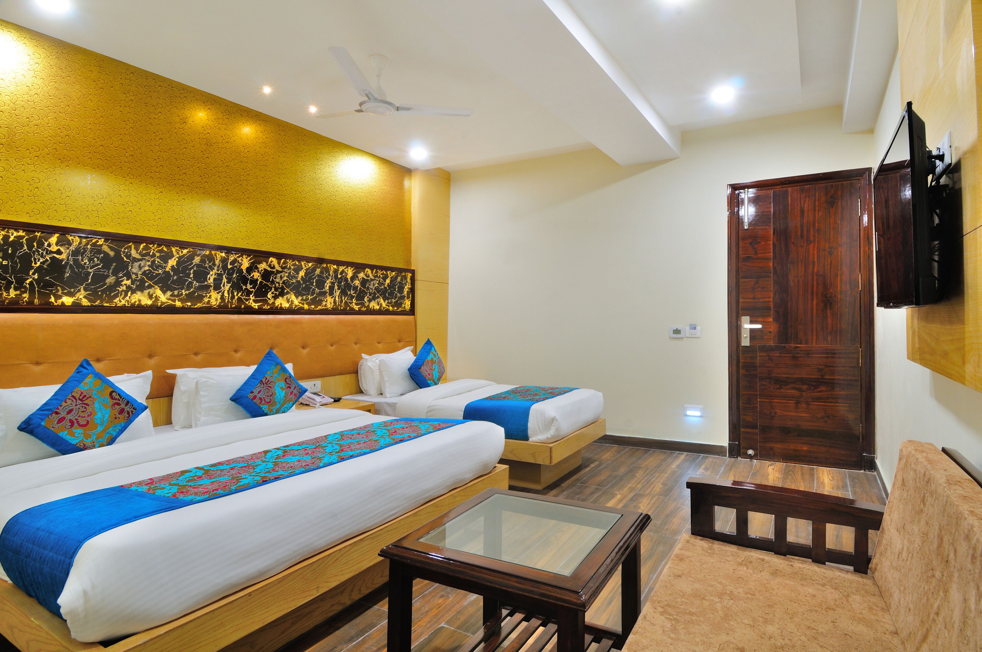 A hotel room with two beds, a wooden bench, a glass-top table, and a television mounted on the wall.