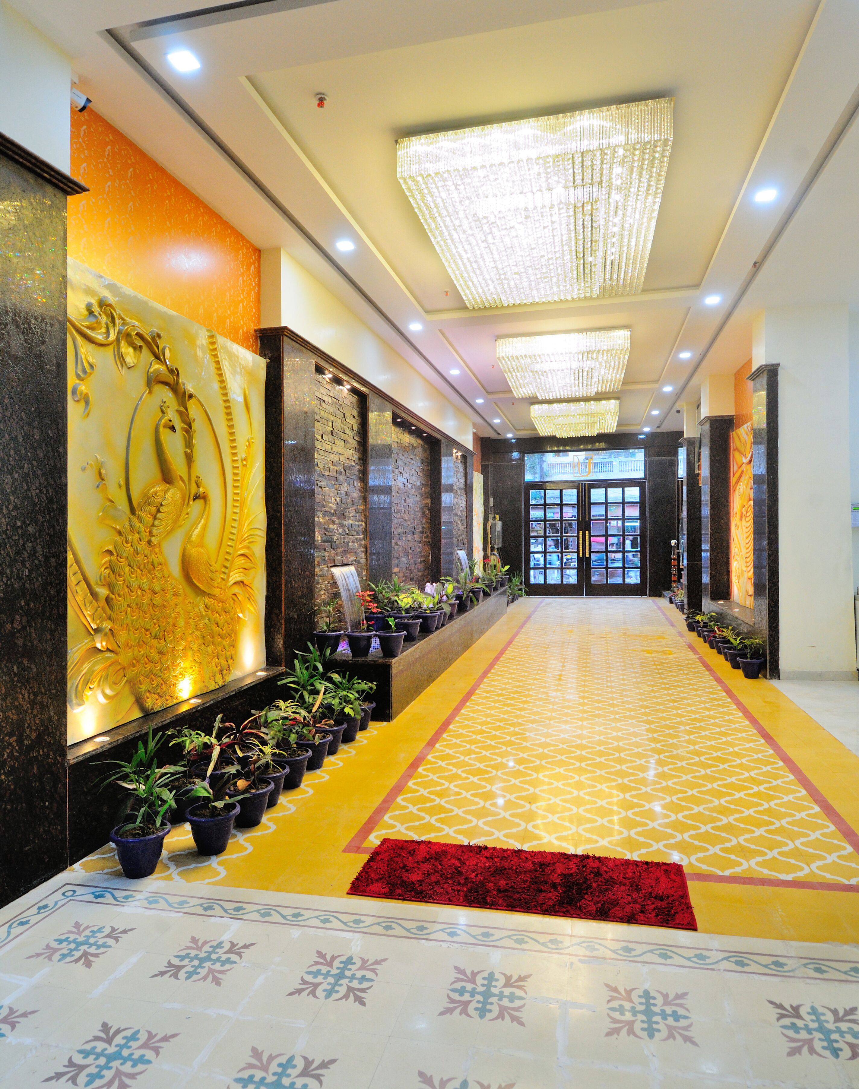 A hotel lobby with a long corridor, decorative wall panels, potted plants, a patterned floor, and a red carpet.