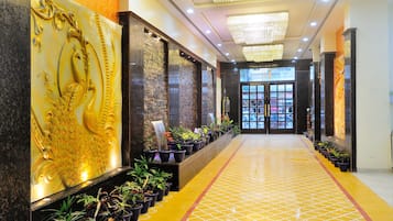 A hotel lobby with a long corridor, decorative wall panels, potted plants, a patterned floor, and a red carpet.