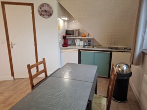 A compact kitchen with a microwave, coffee maker, and a sink. There's a dining table with chairs and a trash bin.