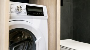 Une machine à laver compacte et moderne, dotée d’un écran numérique et d’un panneau de commande, installée dans une petite salle de bain bien éclairée, avec un meuble-lavabo en bois et des toilettes blanches.