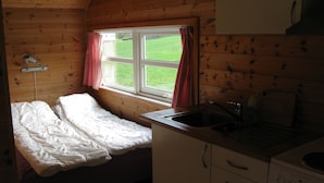 A small, cozy cabin with a single bed, a kitchen area with a sink and stove, and a window with a view of a grassy field. - Sæbø Camping (Eidfjord)