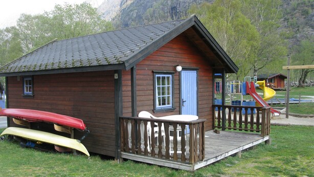 A wooden cabin with a porch, a red and yellow canoe, and a playground with a slide. - Sæbø Camping (Eidfjord)