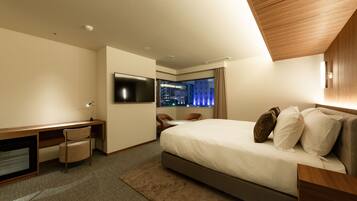 A hotel room with a bed, a desk, a chair, a TV, and a view of the city at night.