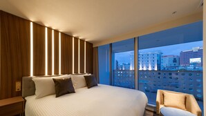 A modern hotel room with a large bed, a seating area with a chair and table, and a city view through large windows. - The OneFive Villa Fukuoka (Fukuoka)
