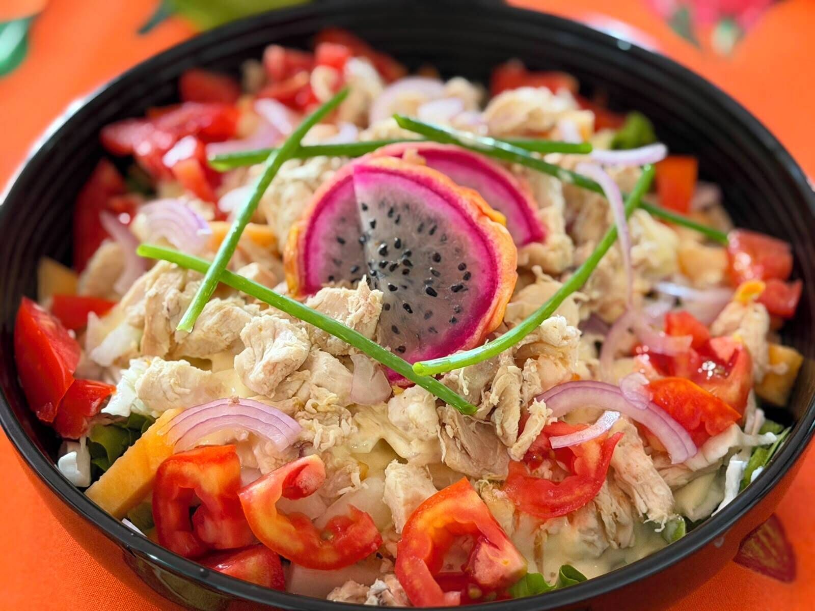 Un tazón de comida con diversos ingredientes, incluyendo pollo, verduras y una porción de pitahaya.