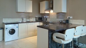 A modern kitchen with white cabinets, a built-in washing machine, a microwave, and a coffee maker. There are two white bar stools at the island. - 59 Riverun (Belturbet)