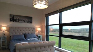 A bedroom with a large bed, a bench, and a view of a grassy field through large windows. - 59 Riverun (Belturbet)