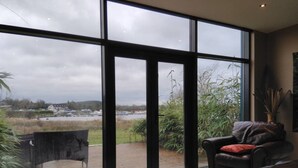 A modern living room with large windows, a leather sofa, and a view of a water body and greenery. - 59 Riverun (Belturbet)
