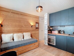 Una habitación acogedora con pared de madera, un sofá con almohadones mullidos, una cocina con armarios oscuros y una lámpara colgante.