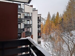 A multi-story building with balconies, a snow-covered ground, and trees with autumn foliage.