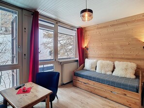 A cozy room with a wooden wall, a sofa with fluffy cushions, a table with a remote control, and a window with red curtains.