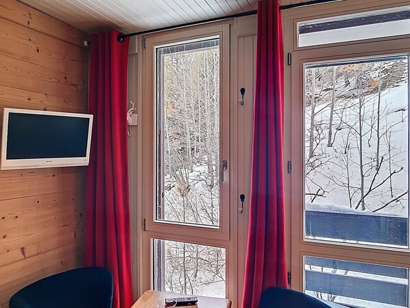 A room with wooden walls, a television mounted on the wall, a window with red curtains, and a view of trees outside.