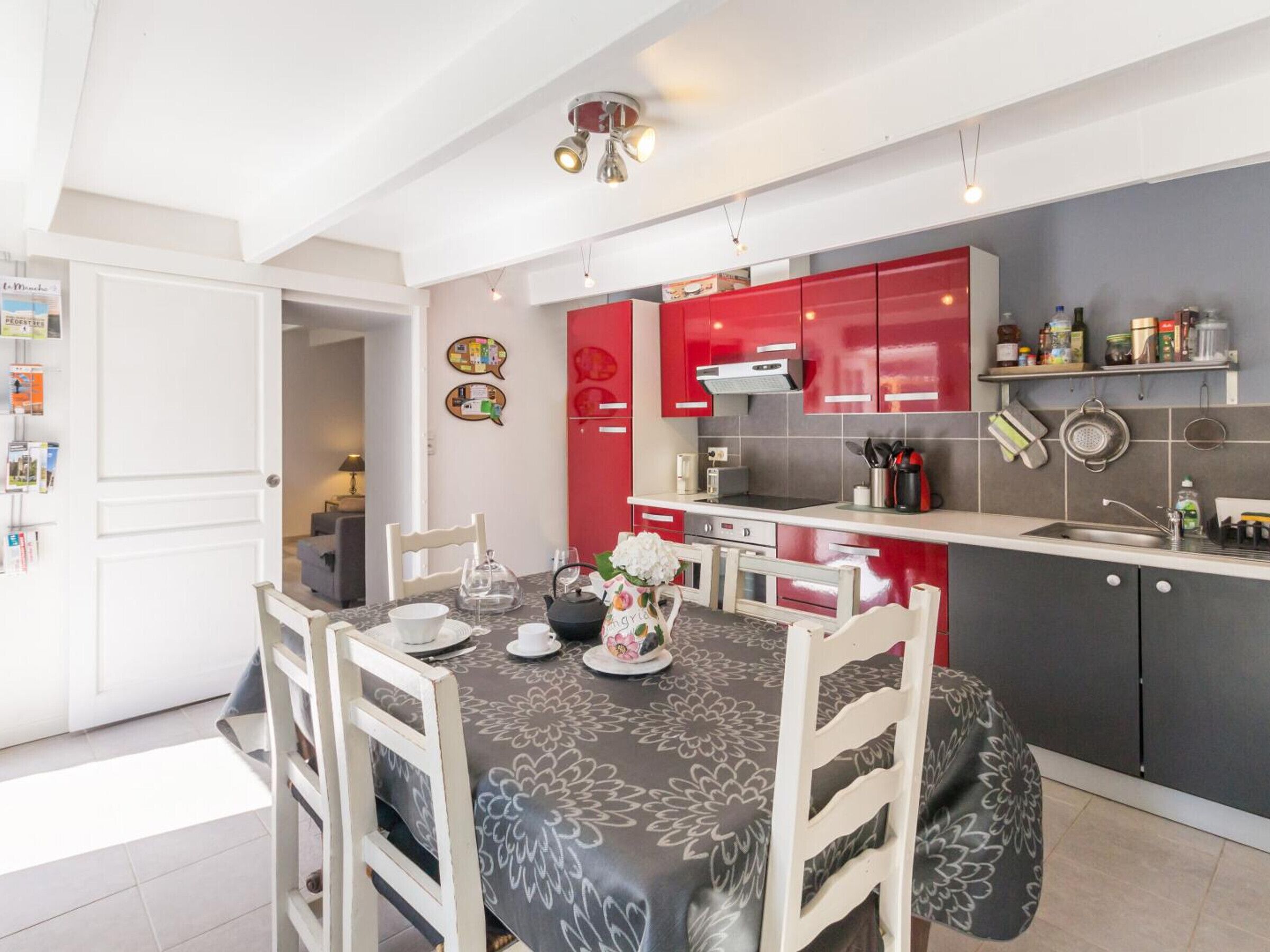 A modern kitchen with red cabinets, a dining table set with white chairs, and a grey patterned tablecloth.