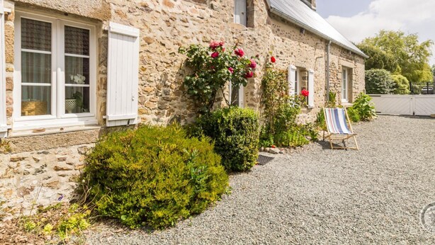 Ein Steinhaus mit weißen Fensterläden, einer Schotterauffahrt und einem kleinen Garten mit Sträuchern und Blumen.
