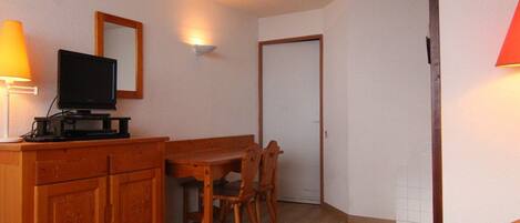 A small, cozy room with a wooden dresser, a television, a dining table with chairs, and a door.