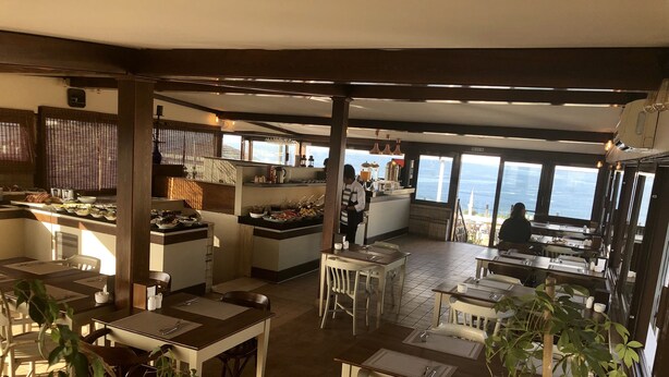 A restaurant with a high ceiling, wooden beams, and large windows offering a view of the sea. Tables are arranged for dining, and a kitchen area is visible in the background. - Mehmet Bey Suites (Istanbul)