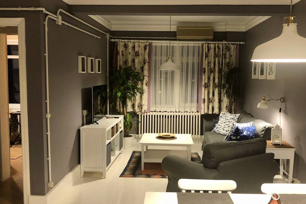 Uma sala de estar moderna com um sofá branco, uma mesa de centro e uma mesinha de cabeceira com abajur. Há um radiador, uma planta em vaso e um espelho na parede.