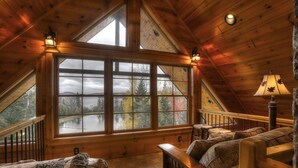 A cozy cabin interior with a large window offering a view of a lake and trees, wooden beams on the ceiling, and a cushioned seating area. - 🐾28 Bunker Ave - Shared waterfront and access to Rangeley Lake, dog friendly (Rangeley)