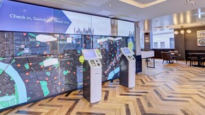A modern hotel lobby with a digital map display, check-in kiosks, and a herringbone wood floor. - hub by Premier Inn London Earl's Court (West Brompton) (London)