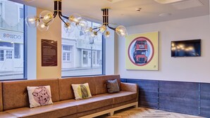 A modern hotel lobby with a wooden bench, a chandelier, a wall-mounted TV, and a large window with a view of the street. - hub by Premier Inn London Earl's Court (West Brompton) (London)