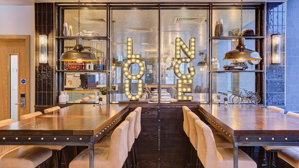 A restaurant with a long wooden table and chairs, a large illuminated sign reading 'Lounge,' and a view through a glass wall. - hub by Premier Inn London Earl's Court (West Brompton) (London)