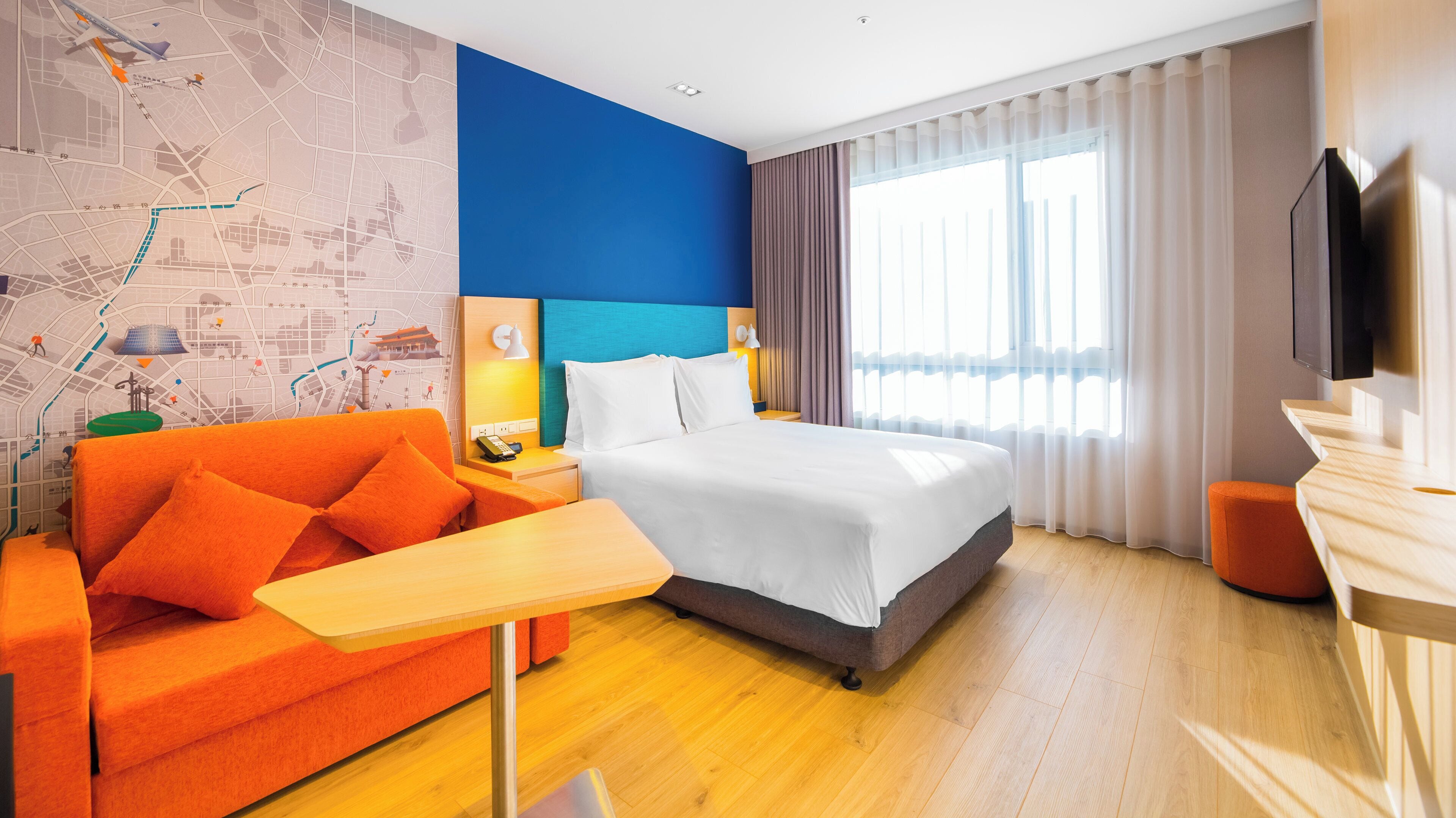 A hotel room with a bed, a desk, a sofa, a map wall, and a TV.