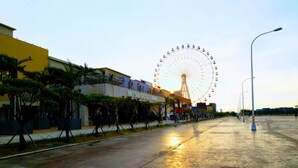 Au loin, on aperçoit une grande roue, un bâtiment portant l’inscription « MITSUI OUTLET PARK », et une promenade pavée bordée d’arbres et de lampadaires.