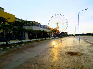 Au loin, on aperçoit une grande roue, un bâtiment portant l’inscription « MITSUI OUTLET PARK », et une promenade pavée bordée d’arbres et de lampadaires.