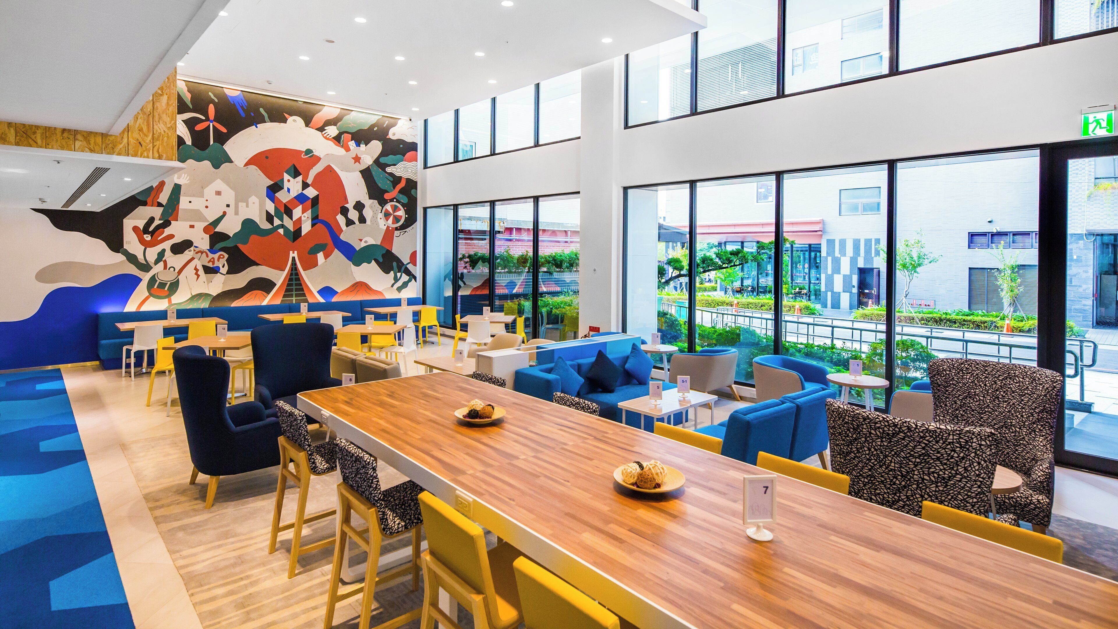 A modern restaurant with a long wooden table, blue and yellow seating, and a colorful mural on the wall.