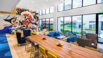 A modern restaurant with a long wooden table, blue and yellow seating, and a colorful mural on the wall.