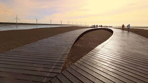 Une promenade en bois menant à une plage, avec des éoliennes en arrière-plan.