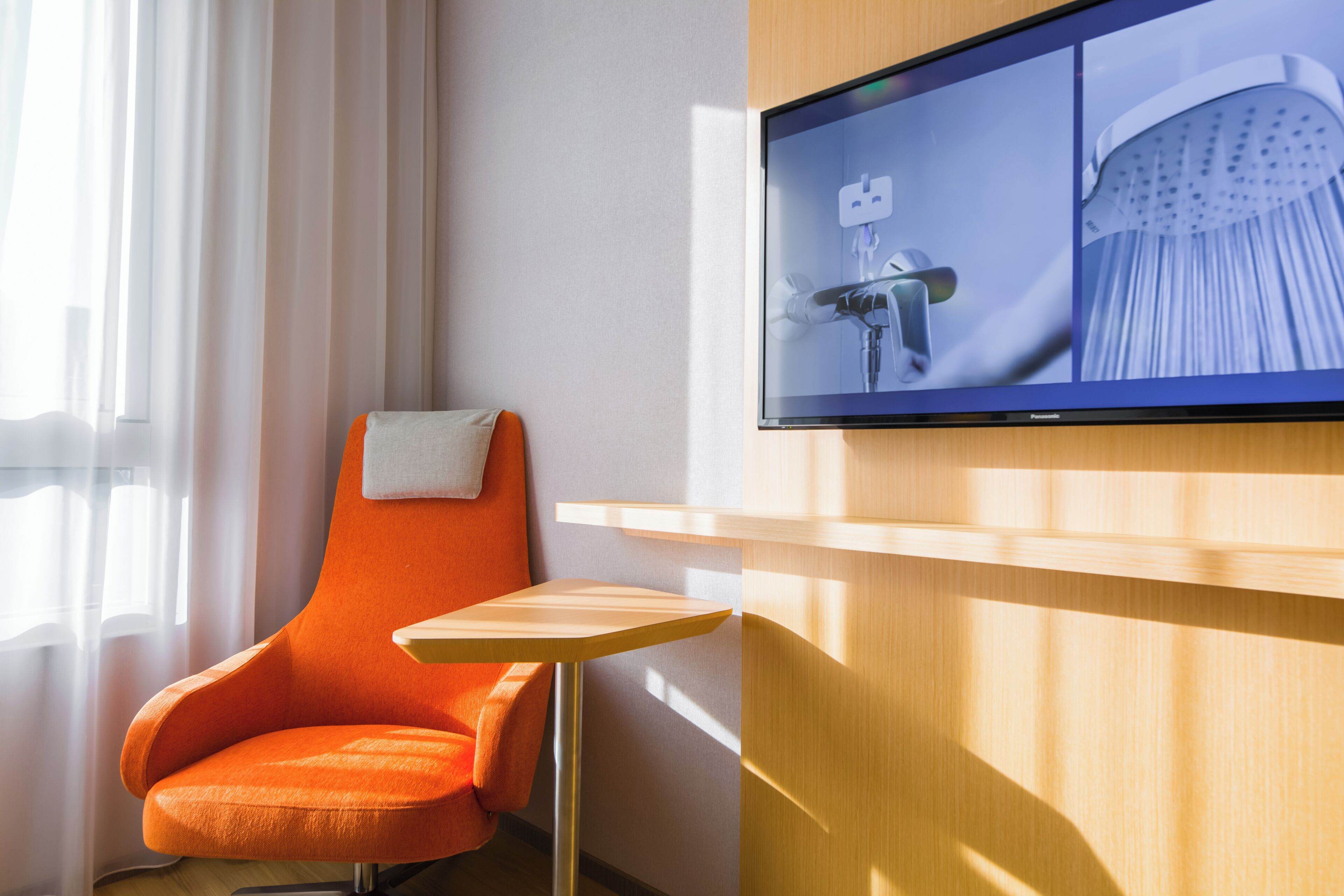 A modern hotel room with a large flat-screen TV mounted on the wall, a wooden side table, an orange armchair, and a window with sheer curtains.