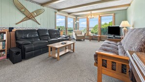 A spacious living room with a black leather sofa, a wooden coffee table, and a grey patterned sofa. There is a television on a stand, a bookshelf, and a large window offering a view of greenery outside. - 🐾Dallas Ridge - Dog friendly four seasons home in Rangeley! (Rangeley)