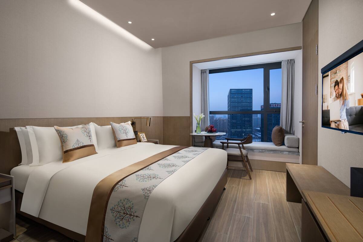 Quarto de hotel moderno com uma cama grande, uma mesa pequena, um sofá e vista para a paisagem urbana através de uma janela ampla.