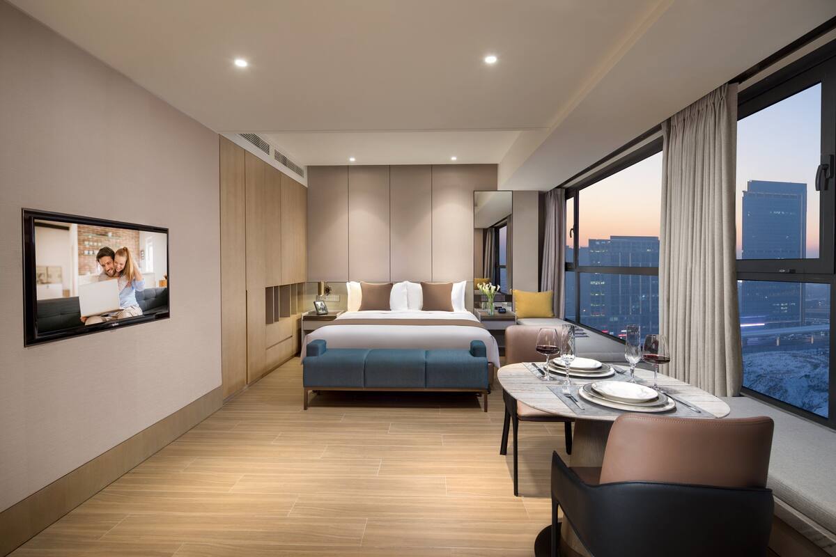 Quarto de hotel moderno com uma cama grande, área de jantar com mesa posta para dois e vista para a cidade através de janelas amplas.