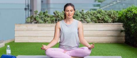 A person sitting cross-legged on a yoga mat in an outdoor area with artificial grass, a water bottle, and a rolled-up mat nearby.