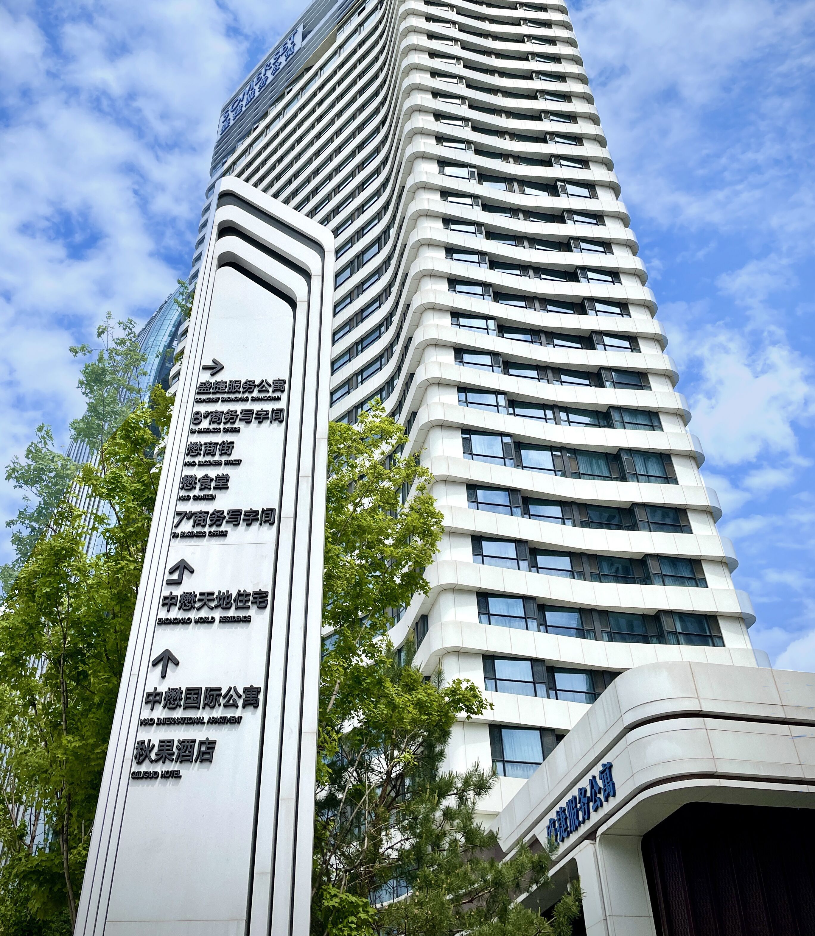 Un edificio alto y moderno con fachada curva y numerosas ventanas. Se observa un gran cartel vertical con flechas direccionales y texto que indica diferentes ubicaciones y servicios.