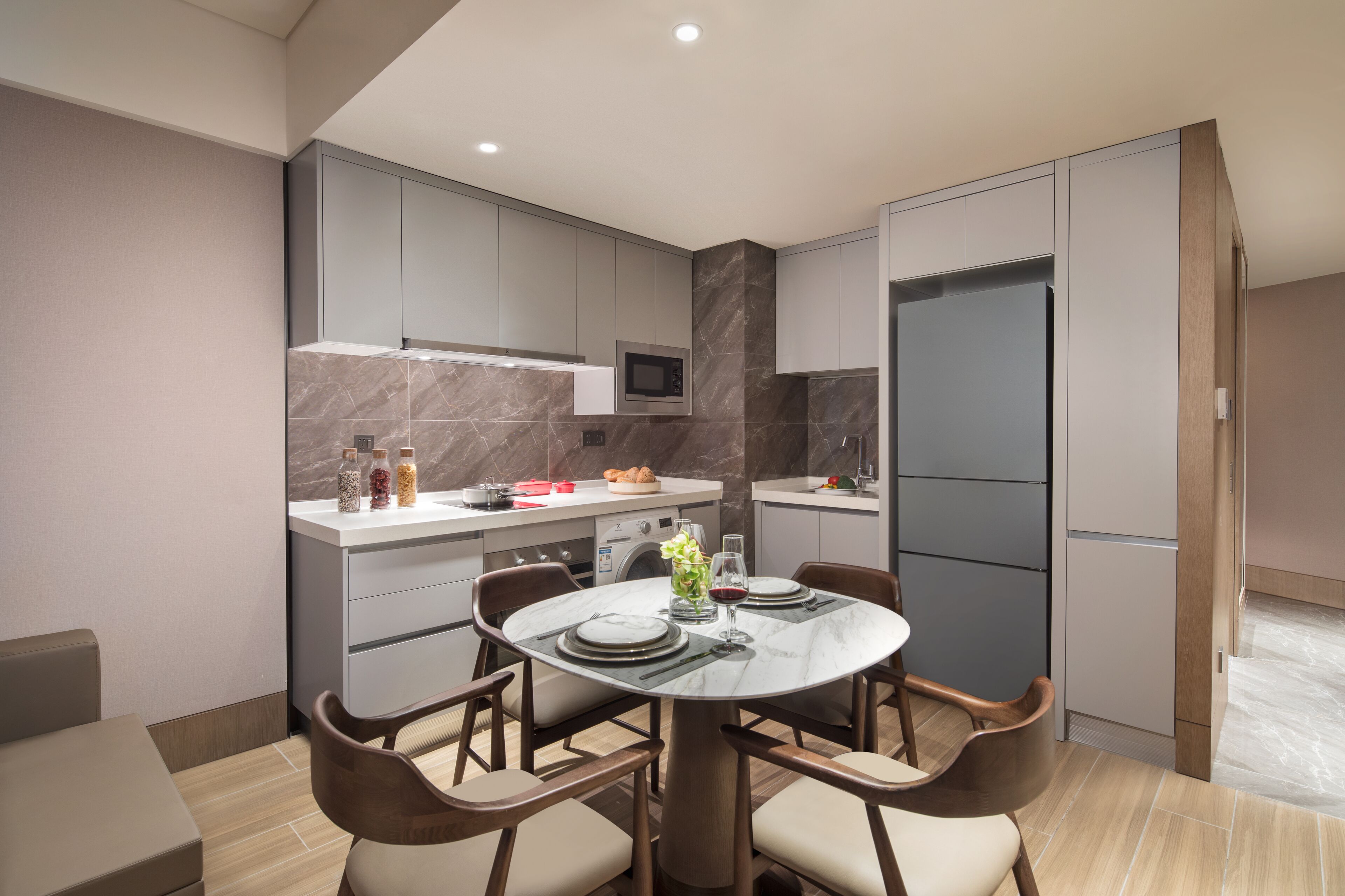 Une cuisine moderne avec des meubles gris, un comptoir en marbre et un micro-ondes encastré. On y trouve une table à manger avec des chaises, ainsi qu’une petite table d’appoint agrémentée d’un vase et de verres à vin.