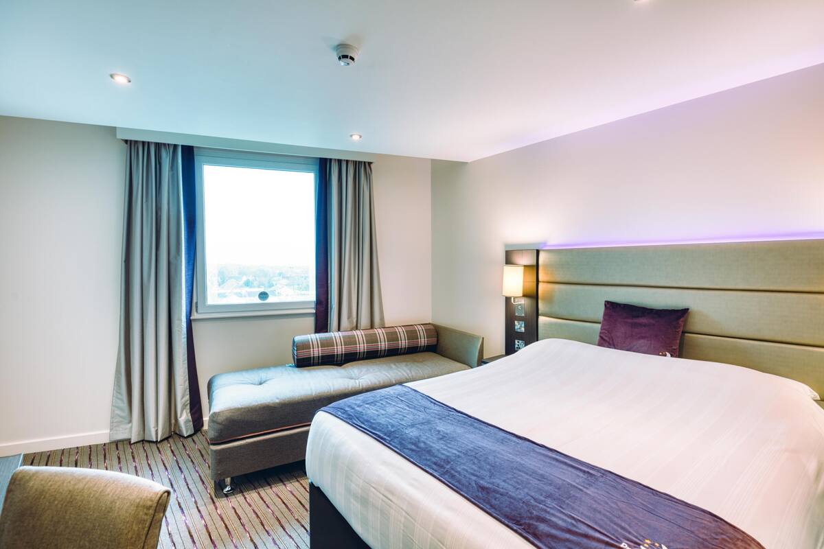 Una habitación de hotel con una cama grande, un sofá más pequeño, una ventana con cortinas, una mesita de noche con lámpara y vistas a la ciudad.