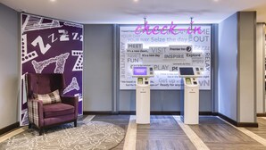 A hotel lobby with a purple wall, a check-in kiosk, a plush armchair, and a patterned rug. - Premier Inn London Sutton (Sutton)