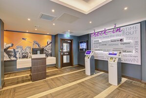 Zona de recepción de hotel con un quiosco de registro digital, mostrador de recepción y una pared con frases motivacionales.
